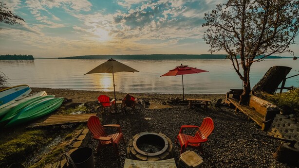 Un espacio de descanso junto al lago con una hoguera, sillas rojas y sombrillas.