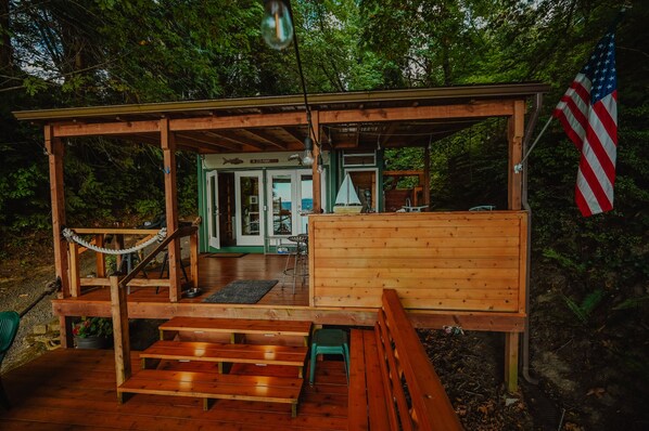 A wooden cabin with a porch, a flag, and a covered area with a bench. - Glamping on the Puget Sound - Small Waterfront Cabin (Olympia)