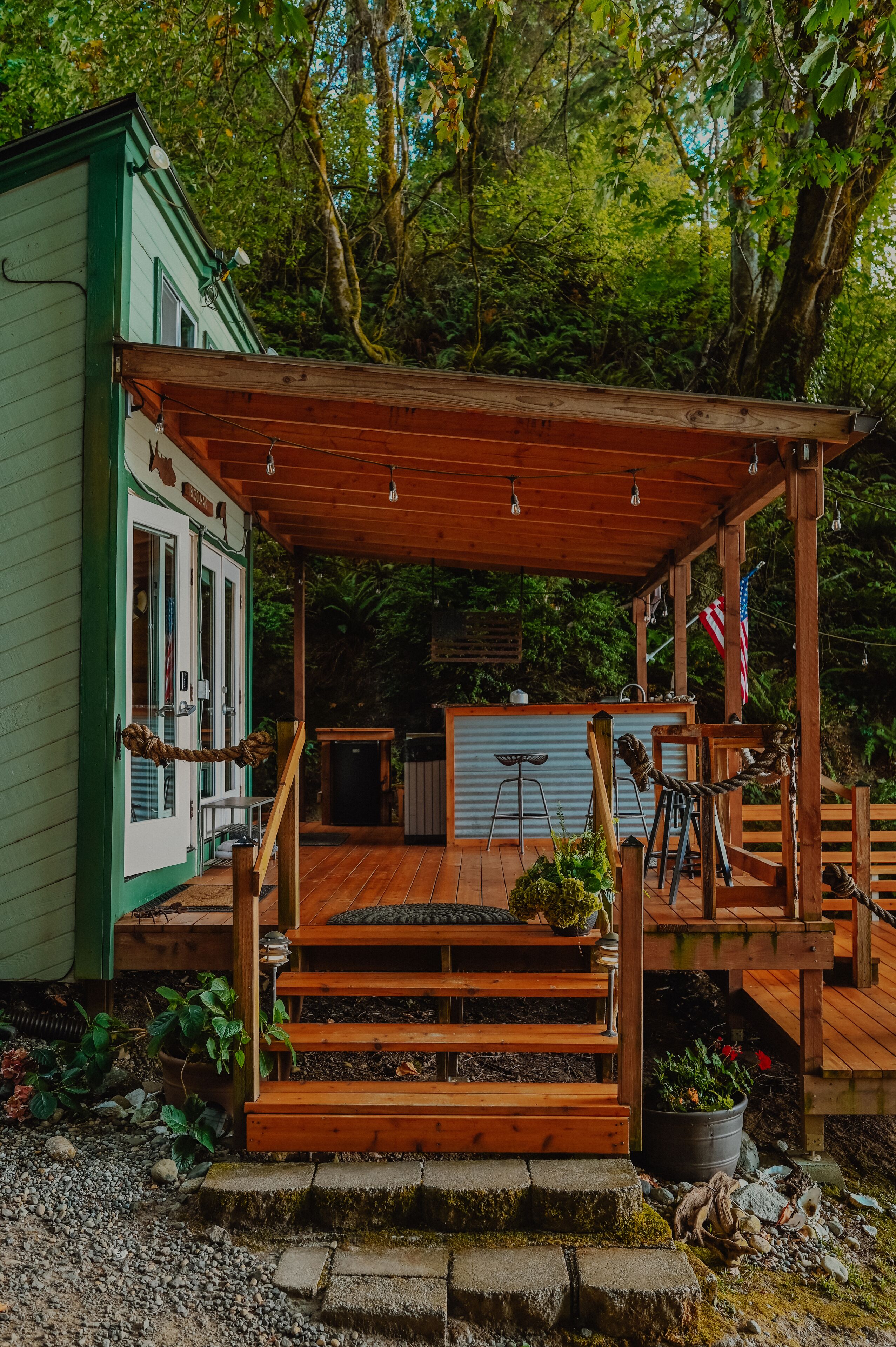 A wooden deck with a pergola, steps leading to a covered area, and a small building with a green exterior.