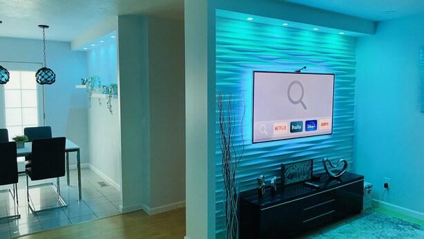A modern living room with a flat-screen TV mounted on a textured wall, a black entertainment unit, and a dining area with a glass table and chairs. - Wendover's most comfortable house for rent. (West Wendover)