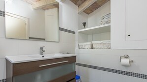 A bathroom with a white sink, mirror, and tiled walls. There are shelves with folded towels and a trash bin. - Villa Les Oliveres Petites en Pollensa by Mallorca North Villas (Pollensa)