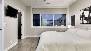 A bedroom with a large bed, a ceiling fan, a TV mounted on the wall, and a window with a view of buildings outside.