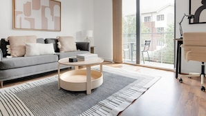 A modern living room with a grey sofa, a round coffee table, and a large window with a view of buildings. - Hip Islington 2BR w/ Day Porter & Balcony, nr Kings Cross, by Blueground (London)