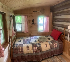 Ein Schlafzimmer in einer Blockhütte mit einer Karo-Decke, zwei Fenstern mit Vorhängen und einem hölzernen Kopfteil.