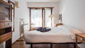 A bedroom with a large bed, wooden headboard, a radiator, a window with curtains, a chair, and a small table with a lamp. - Aiguamoig by SeaMount Rentals (Gessa)