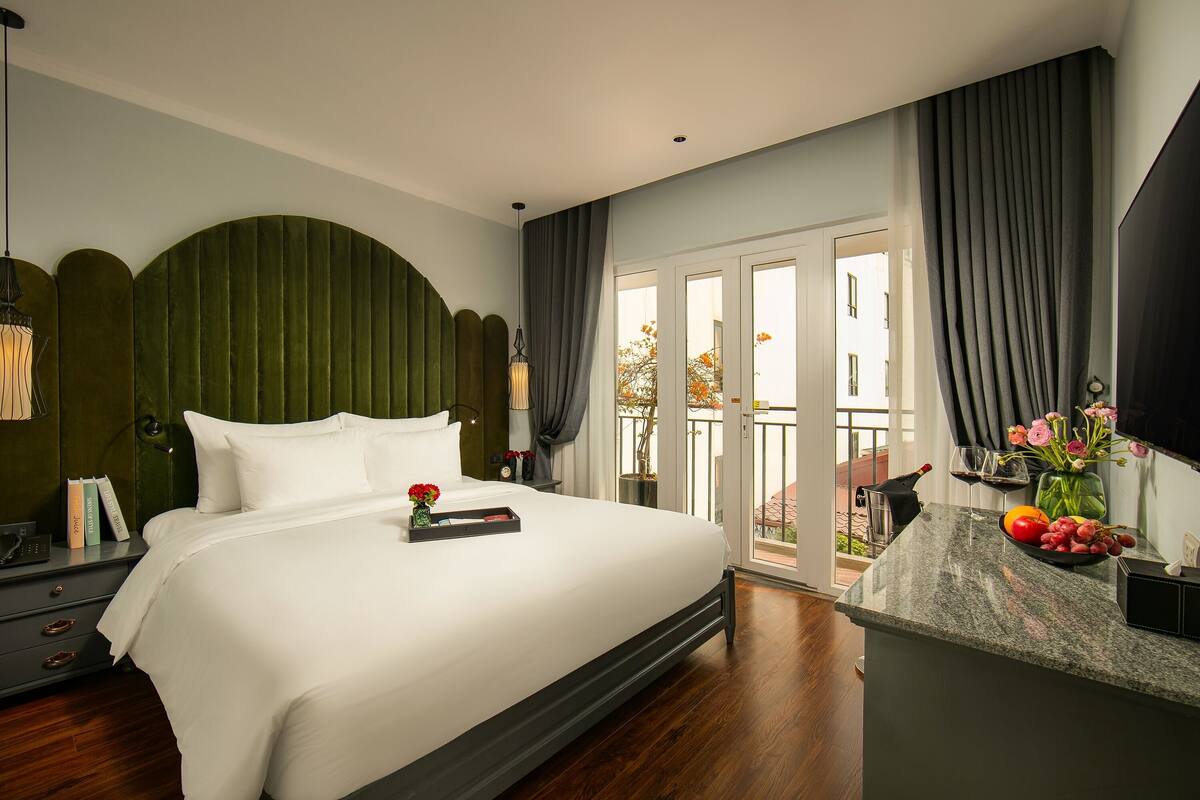 A modern hotel room with a large bed, a nightstand with books, a desk with a vase of flowers, and a view of the outdoors through a sliding door.