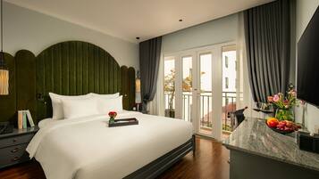 A modern hotel room with a large bed, a nightstand with books, a desk with a vase of flowers, and a view of the outdoors through a sliding door.