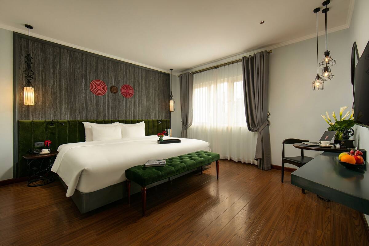 A hotel room with a large bed, a green bench, a desk with a chair, a TV, and a window with curtains.