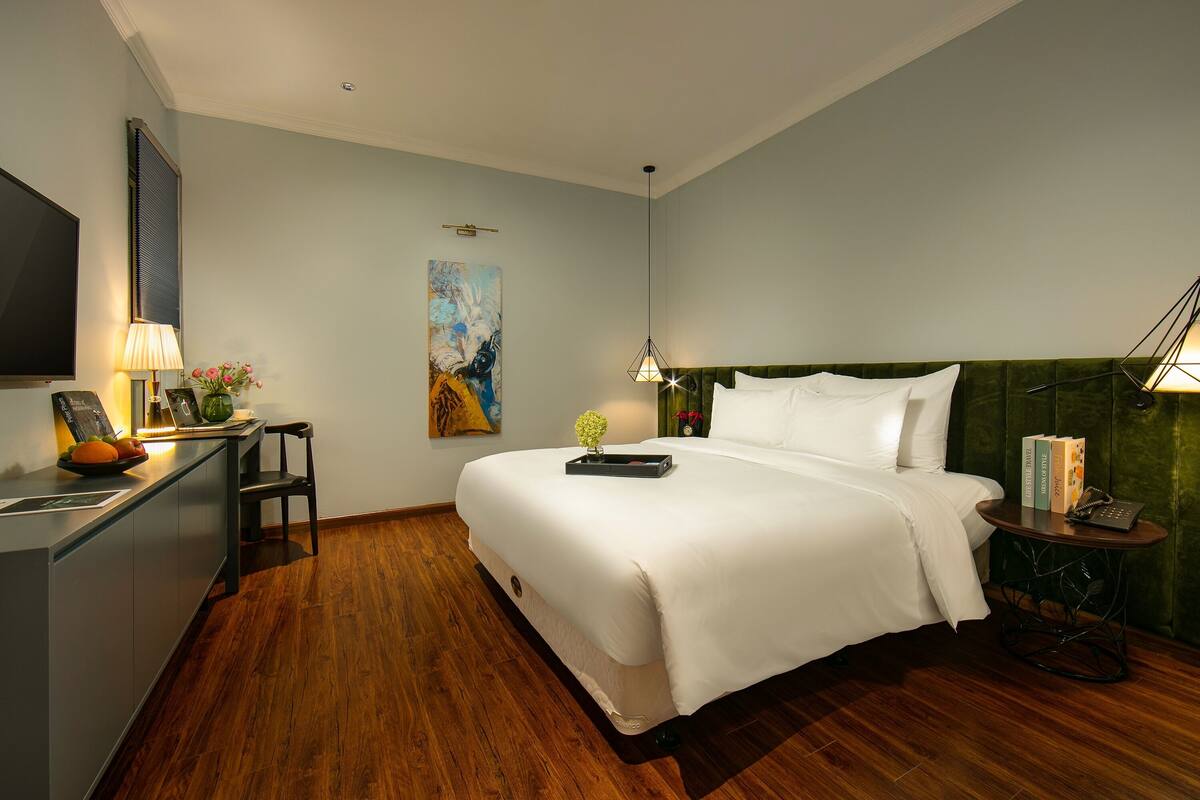 A modern hotel room with a large bed, a desk with a lamp, a chair, and a small table with books.