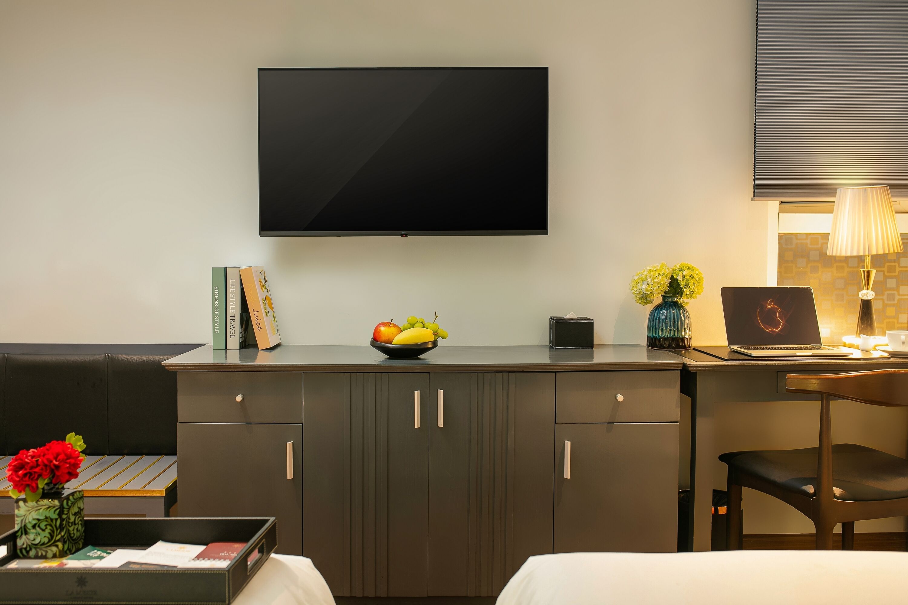 A modern hotel room with a flat-screen TV mounted on the wall, a desk with a laptop, a bowl of fruit, and a vase with flowers.