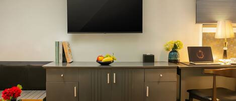 A modern hotel room with a flat-screen TV mounted on the wall, a desk with a laptop, a bowl of fruit, and a vase with flowers.