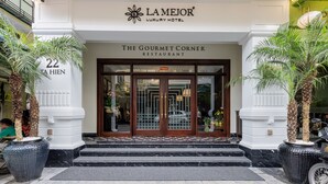 The entrance to Lamejor Luxury Hotel, featuring a grand double door with glass panels, a sign for The Gourmet Corner Restaurant, and a palm tree planter on either side.