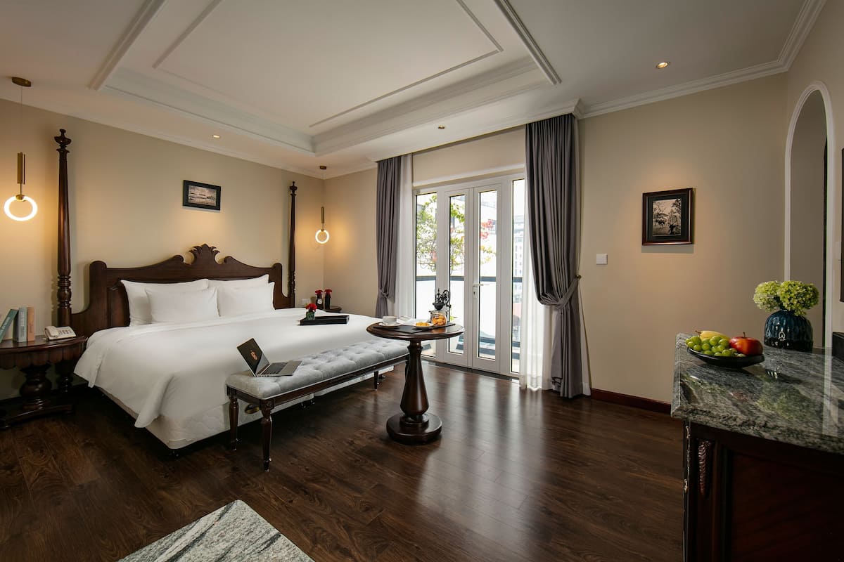 A spacious hotel room with a large bed, a round table with a laptop, a small stool, and a fruit bowl on the counter.