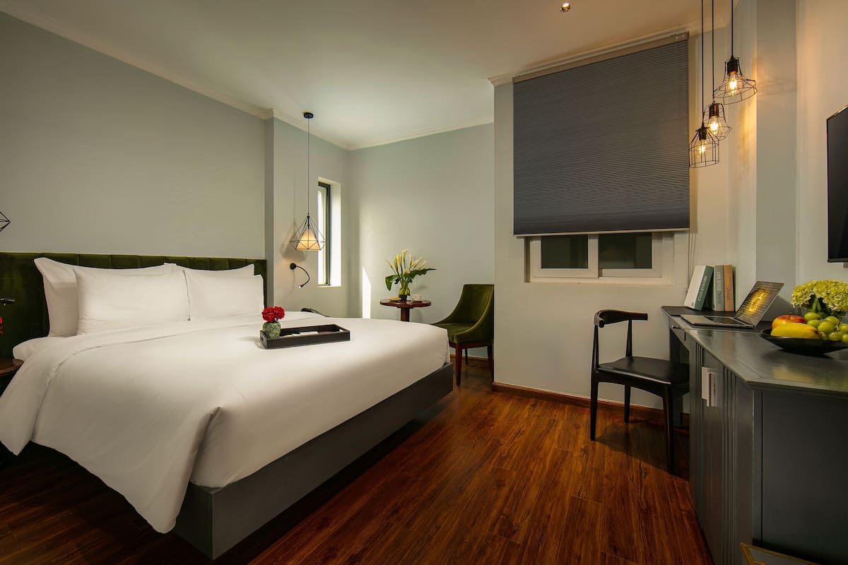 A modern hotel room with a large bed, a desk with a laptop, a chair, and a small table with flowers.