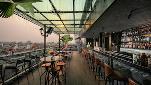 A rooftop bar with a view of the city, featuring tables and chairs, a bar counter, and a variety of bottles.