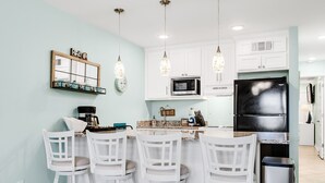 Cucina con mobili bianchi, un frigorifero nero, un forno a microonde e tre sgabelli bianchi.