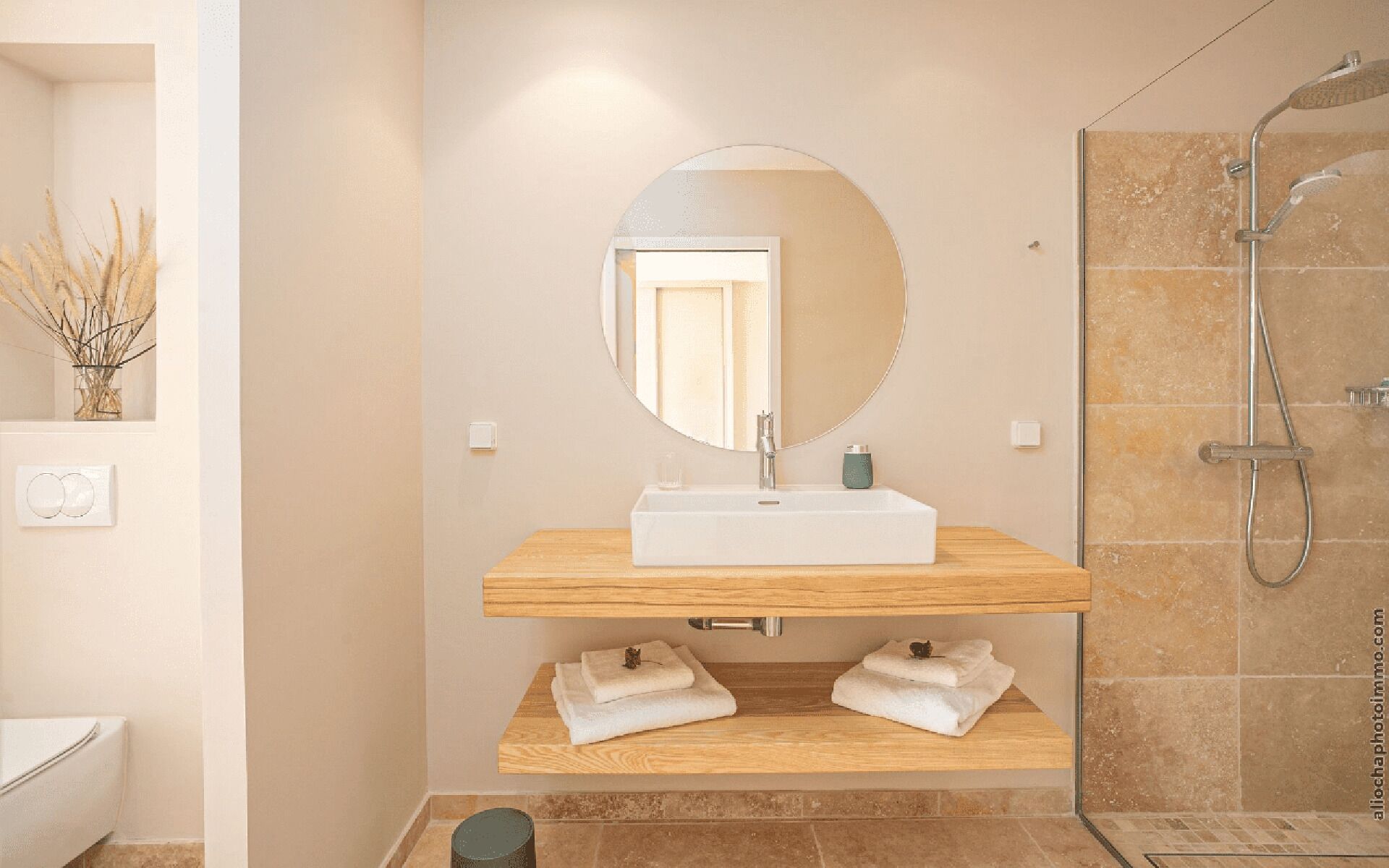 A modern bathroom with a wooden vanity, a round mirror, a shower, and a small shelf with towels.