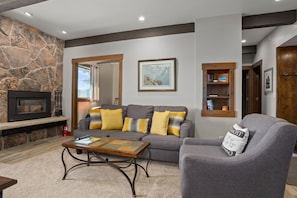 A living room with a stone fireplace, a grey sofa with yellow cushions, a wooden coffee table, and a bookshelf. - Balcony VIEWS - Remodeled - Ski In/Out, Pool, Hot Tub (Steamboat Springs)