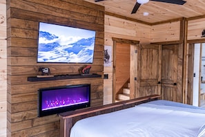 A wooden cabin interior with a bed, a mounted TV, and a fireplace.