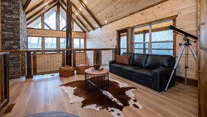 A spacious cabin interior with a high ceiling, wooden walls, large windows, a leather sofa, a round coffee table, and a telescope.