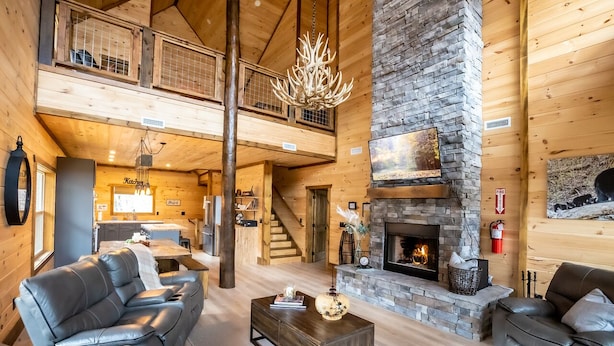 A spacious log cabin interior with a fireplace, leather sofas, a dining area, and a staircase leading to a loft.