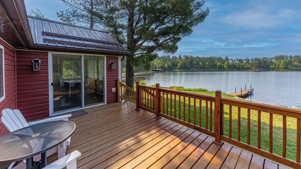 A wooden deck with a table and chairs, overlooking a lake with a dock and a red building with glass doors. - Yacht Club cabin on Burntside, with sauna (Ely)