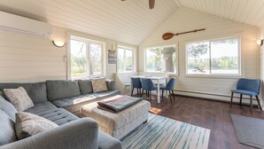 A spacious living area with a large gray sectional sofa, a small ottoman, and a dining table with chairs. There is a ceiling fan, large windows, and a view of the outdoors. - Yacht Club cabin on Burntside, with sauna (Ely)