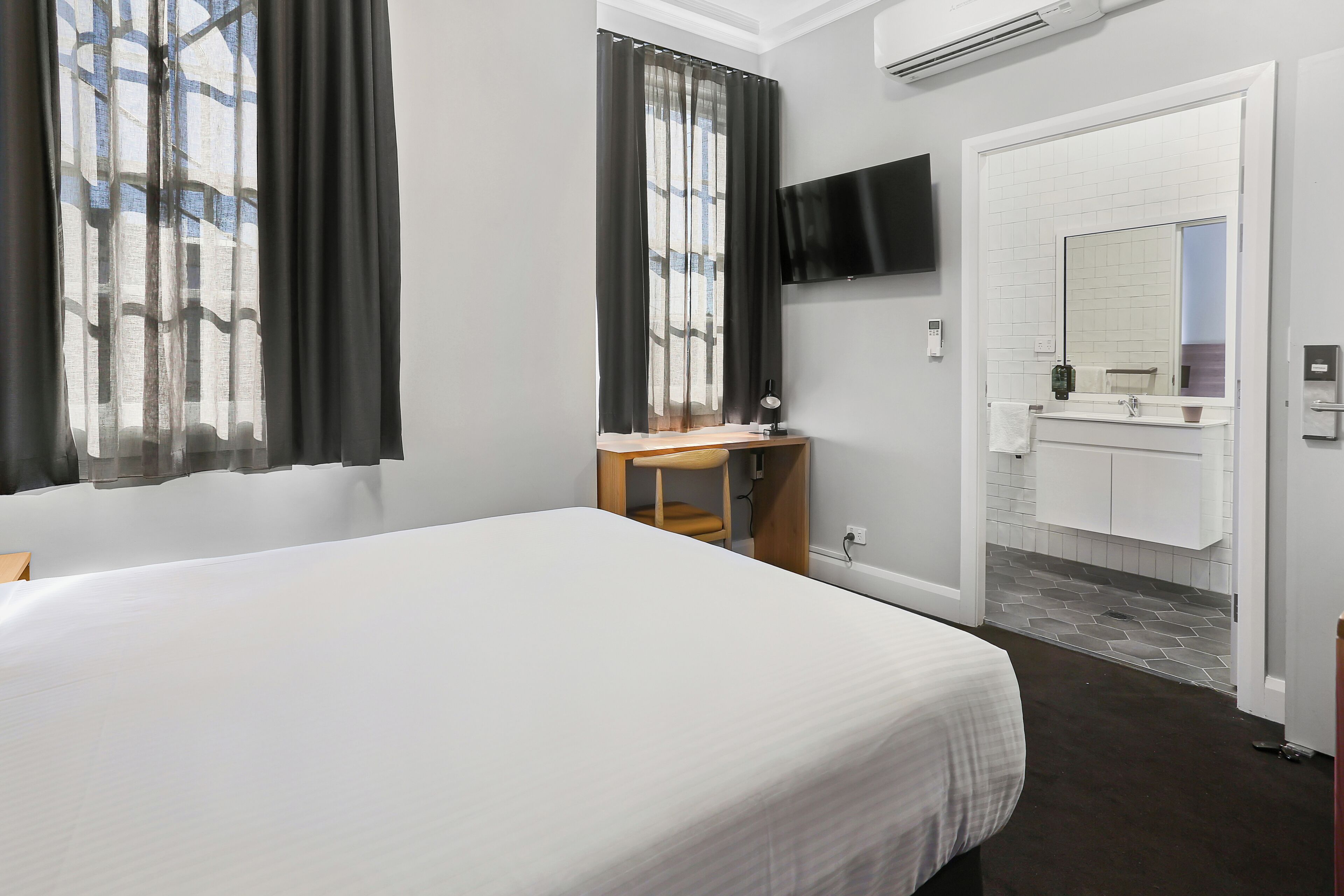A hotel room with a large bed, a desk with a chair, a TV mounted on the wall, a bathroom with a mirror and sink, and a window with curtains.