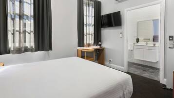 Une chambre d’hôtel avec un grand lit, un bureau avec une chaise, un téléviseur fixé au mur, une salle de bain avec un miroir et un lavabo, et une fenêtre avec des rideaux.