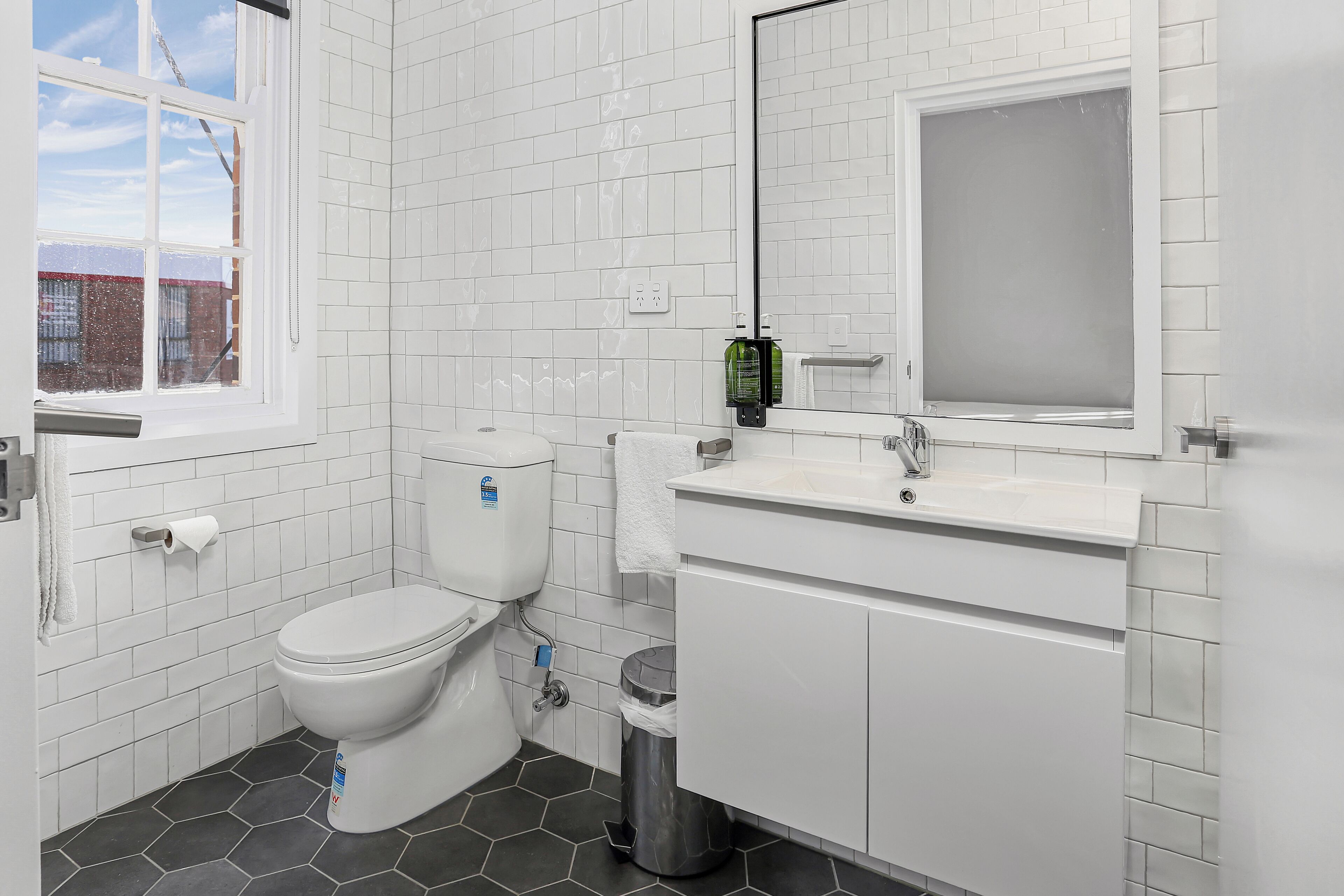 A bathroom with white tiled walls, a white toilet, a sink with a mirror, and a trash can.