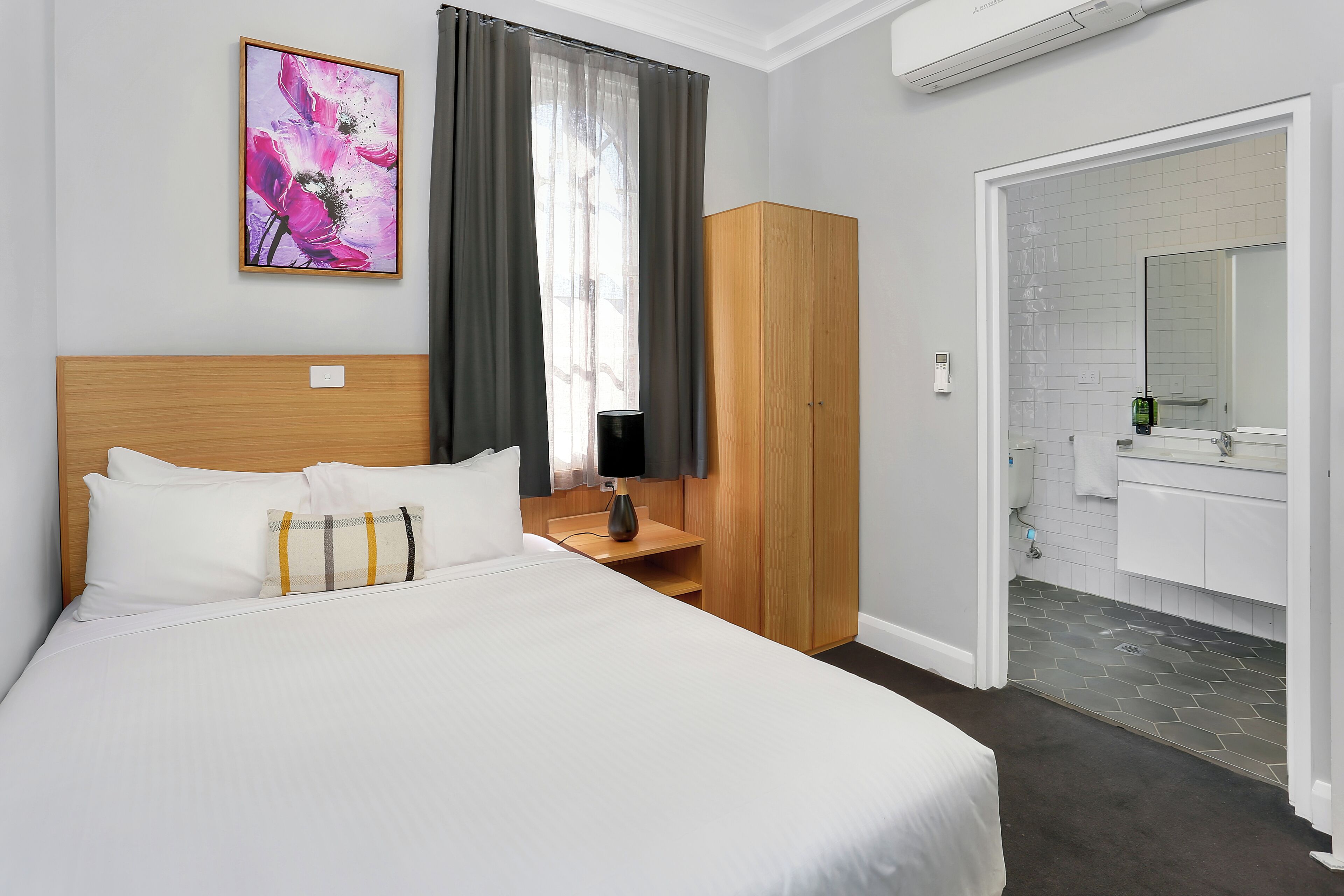 A hotel room with a large bed, a wooden wardrobe, a bedside table with a lamp, a framed artwork on the wall, and a bathroom visible through an open door.