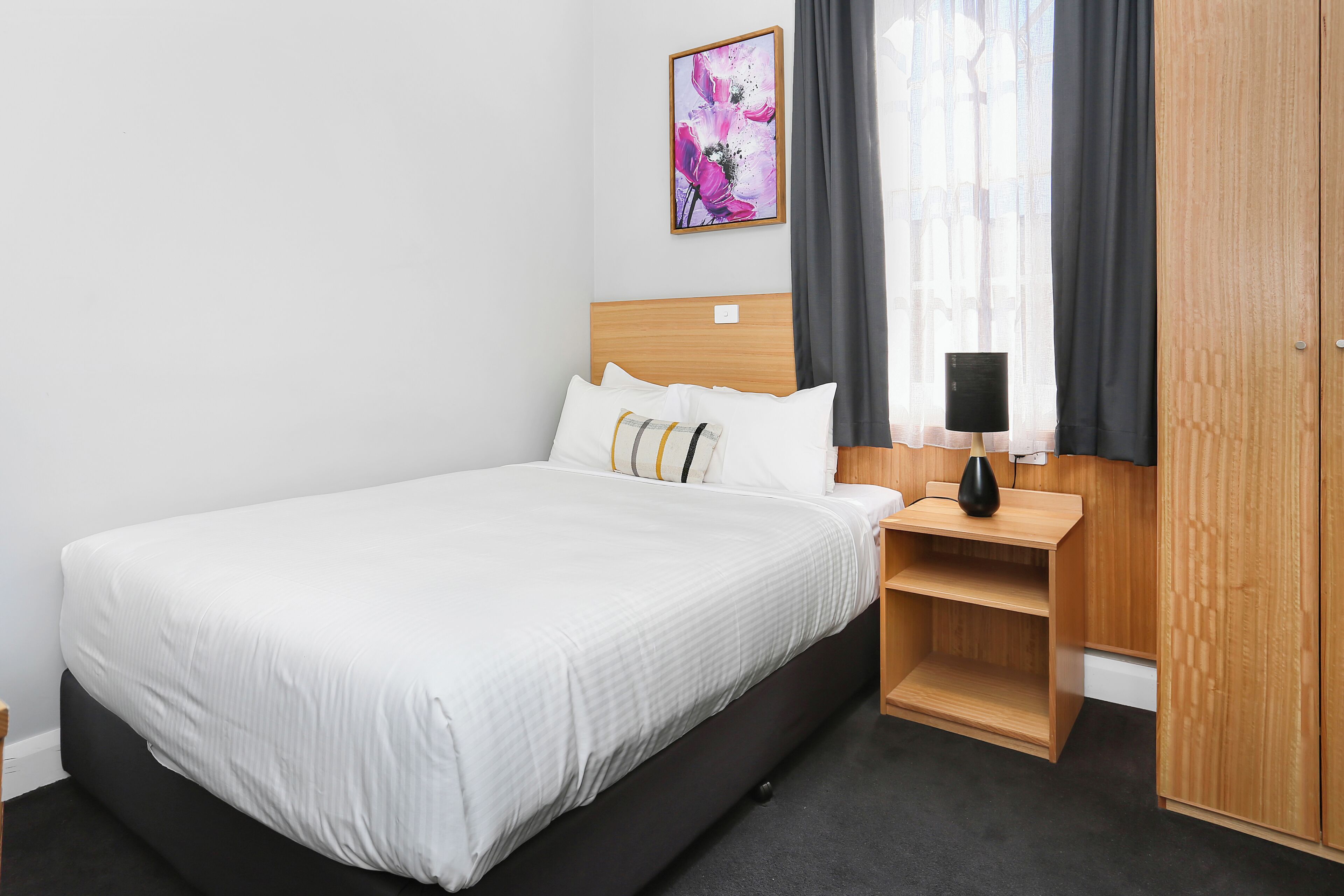 A hotel room with a bed, a nightstand, a lamp, a wardrobe, and a window with curtains.