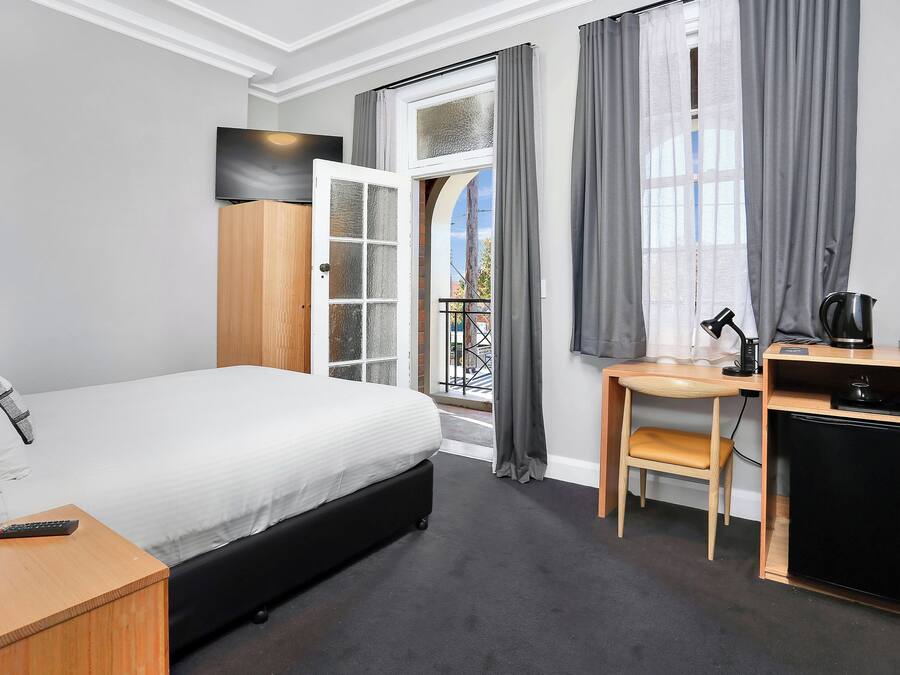 A hotel room with a bed, a desk with a chair, a small refrigerator, a TV, and a window with curtains.