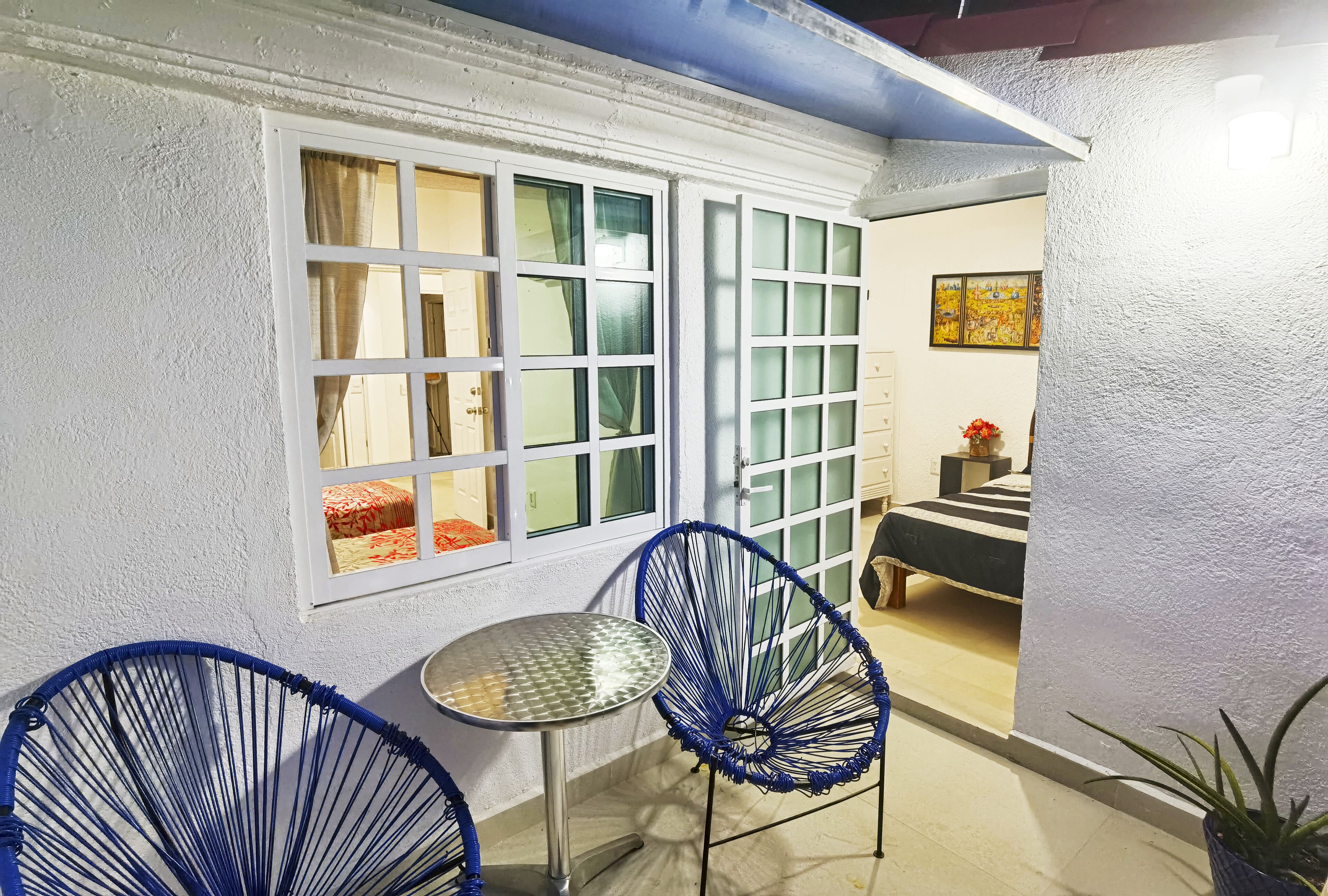 A small room with a white wall, a window with curtains, a metal table, two blue chairs, and a bed with a striped bedspread.