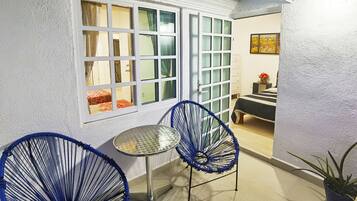 A small room with a white wall, a window with curtains, a metal table, two blue chairs, and a bed with a striped bedspread.