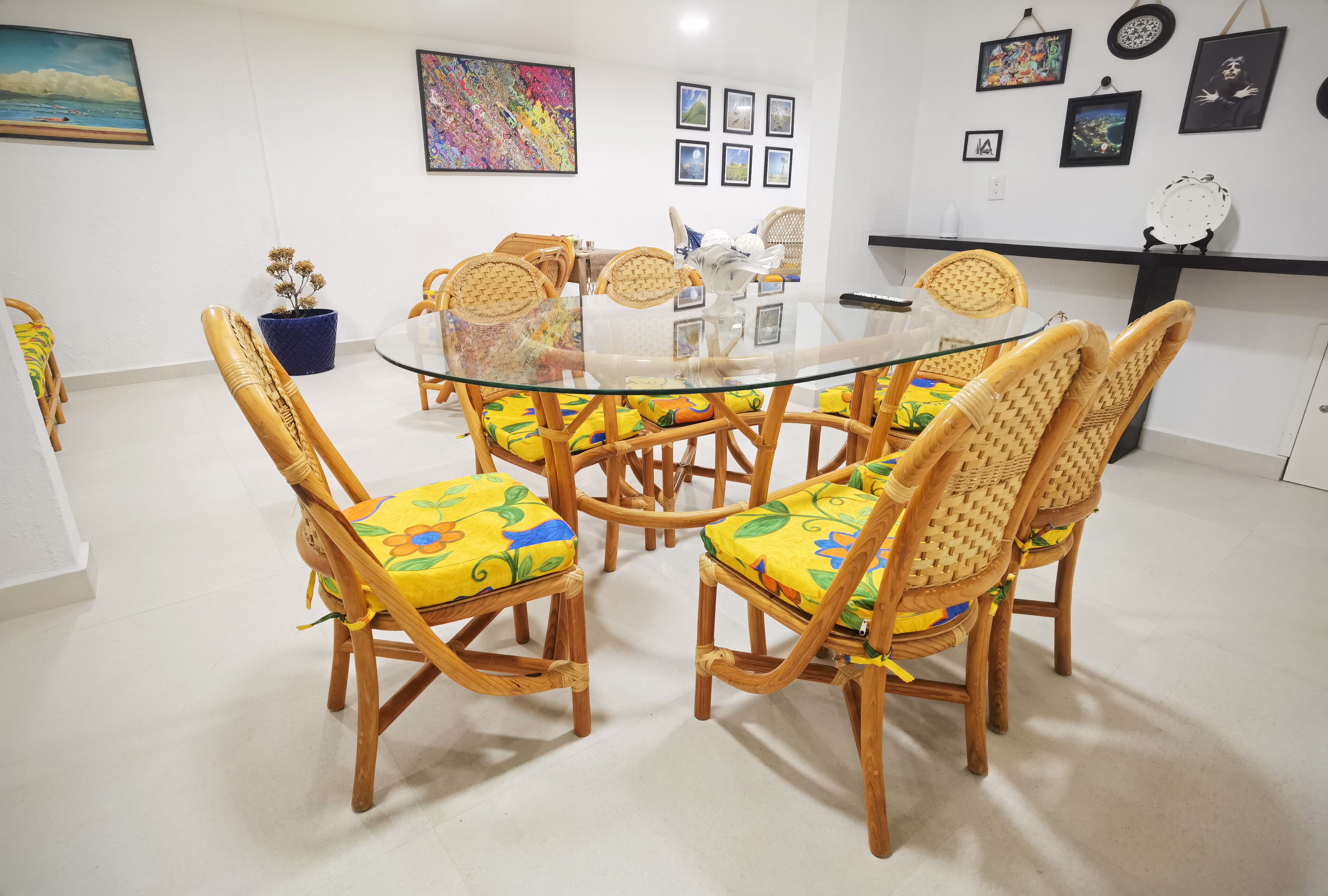 Uma sala de jantar com mesa de vidro, cercada por cadeiras de vime e almofadas coloridas.