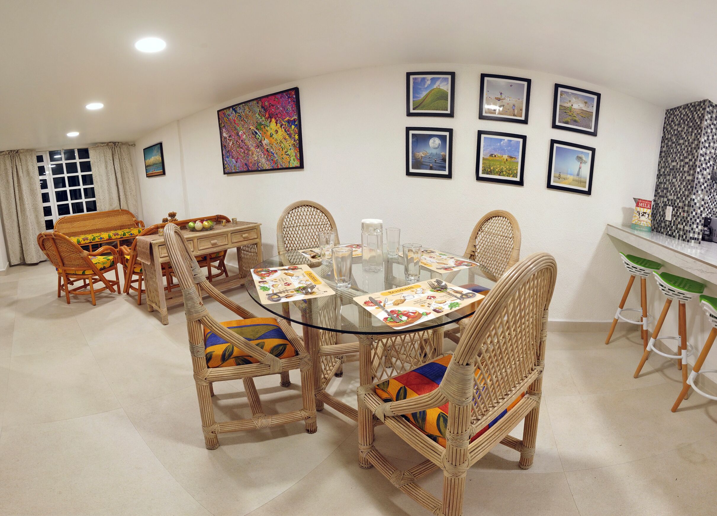 A dining area with a round glass table surrounded by wicker chairs with colorful cushions, a white wall with framed pictures, and a window with curtains.