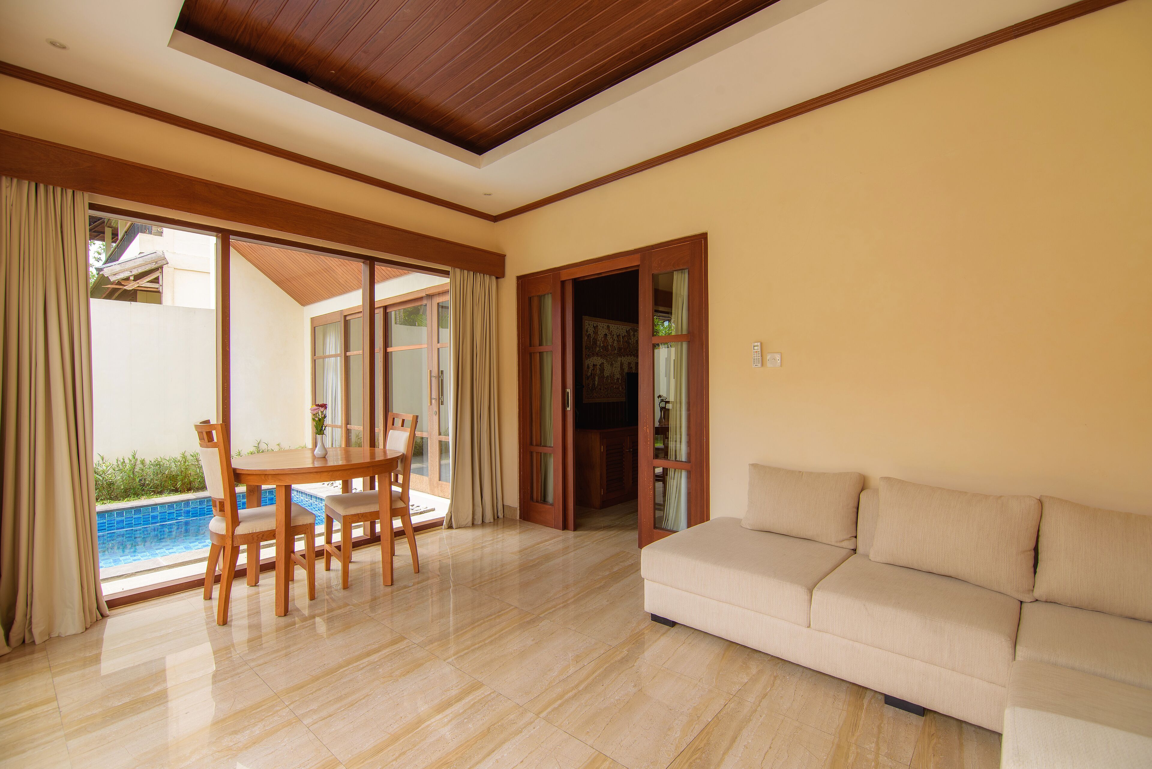 A spacious living room with a beige sofa, a wooden dining table with chairs, and a glass door leading to a pool area.