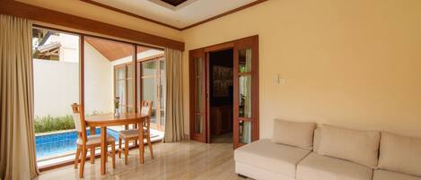 A spacious living room with a beige sofa, a wooden dining table with chairs, and a glass door leading to a pool area.