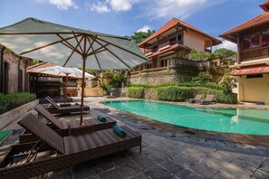 Piscina com cadeiras de descanso e guarda-sóis, cercada por paredes de pedra e vegetação.