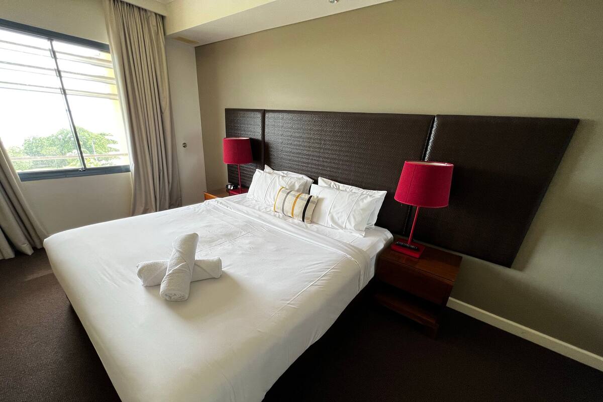 Une chambre d’hôtel avec un grand lit, deux tables de chevet, une tête de lit et une fenêtre avec des rideaux.