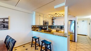 Una cocina moderna con armarios blancos, encimera de granito, refrigerador de acero inoxidable y una zona de comedor con mesa y dos taburetes de bar.