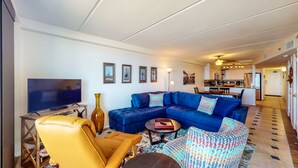 Una sala de estar con un sofá modular, un sillón amarillo, un televisor sobre un soporte de madera y un comedor al fondo.