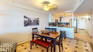 Un coin repas avec une table et des chaises en bois, une cuisine à l’arrière-plan et un ventilateur de plafond.