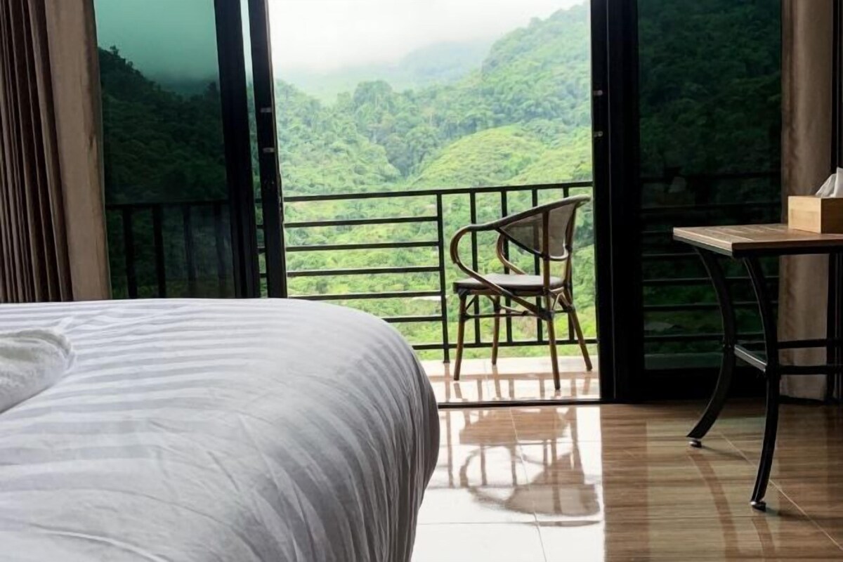 A hotel room with a bed, a chair, a table, and a balcony with a view of a lush green landscape.