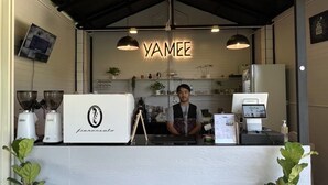 Una zona de recepció amb un cartell de Yamee, una taula d'atenció amb un logotip, una màquina de cafè i dues plantes en testos.