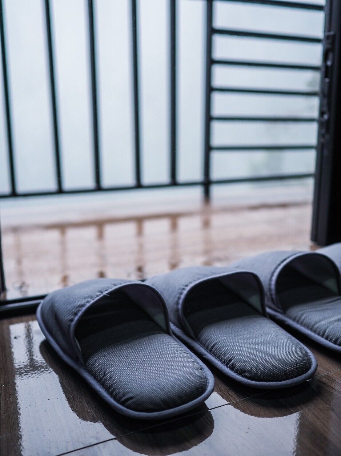 A pair of grey slippers on a wooden floor near a window with a metal railing.