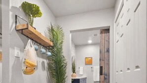 A hallway with wooden flooring, a potted plant, a hanging net with fruits, and a wooden shelf with cups.