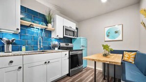 A modern kitchen with white cabinets, a blue tiled backsplash, a microwave, and a stove. There's a small dining table with a plant and a blue sofa with an orange pillow.