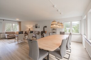 A modern living room with a large wooden dining table, grey chairs, a sofa, and a large window with a view of trees.